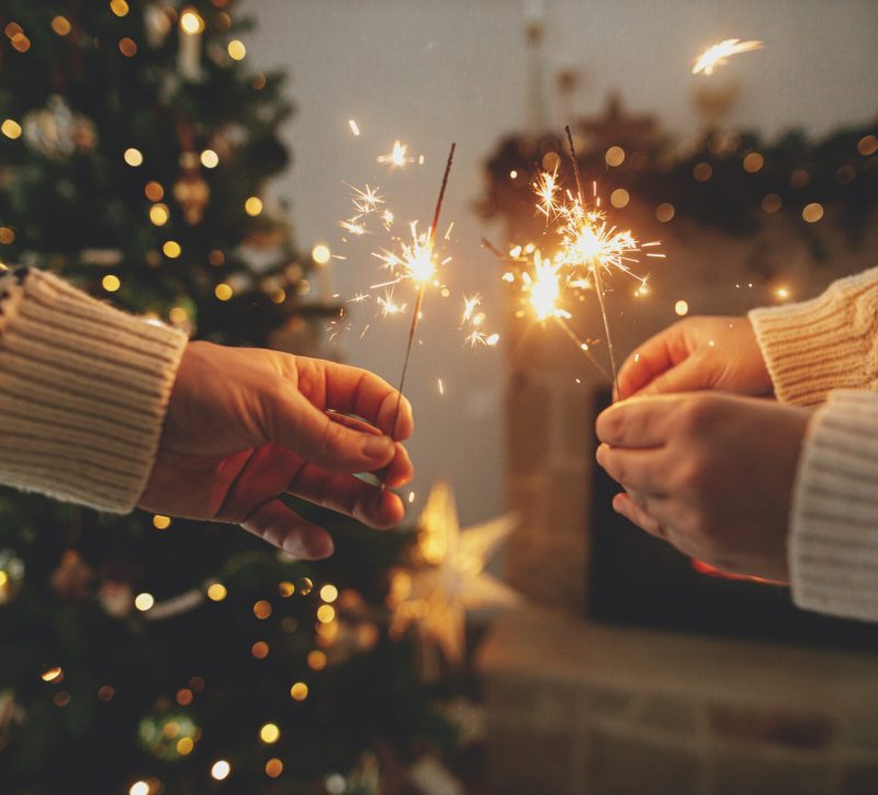 Happy New Year! Hands holding burning fireworks against modern fireplace and christmas tree with golden lights. Friends and family celebrating with burning sparklers in hands, atmospheric eve